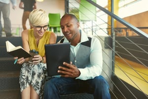 Two creative millenial small business owners working on social media strategy using a digital tablet while sitting in staircase